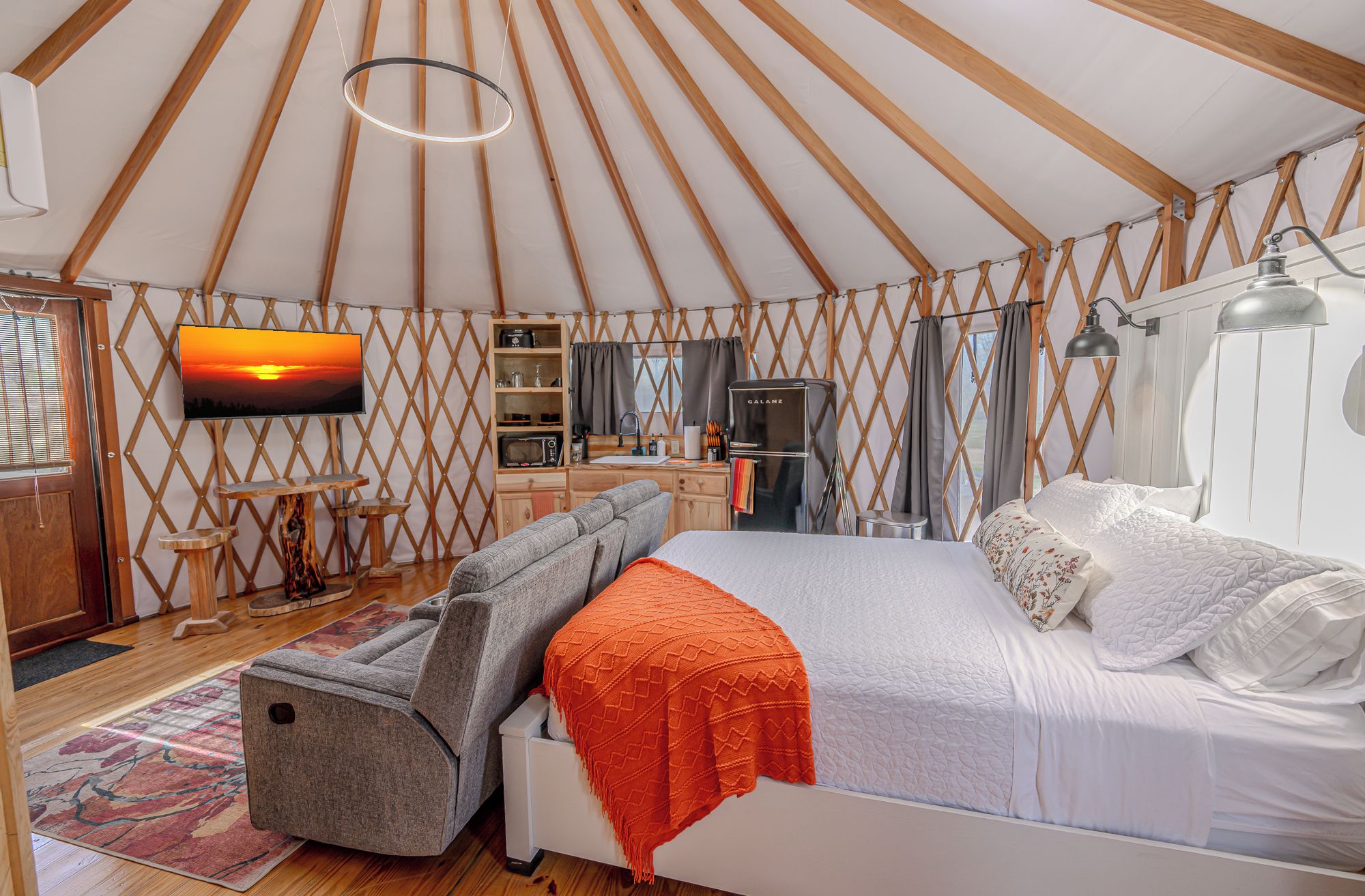 A cozy interior of a yurt with a bed, a TV, a kitchen area, and a dining table.