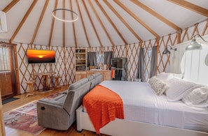 Interior acogedor de una yurta con una cama, un televisor, una zona de cocina y una mesa de comedor.