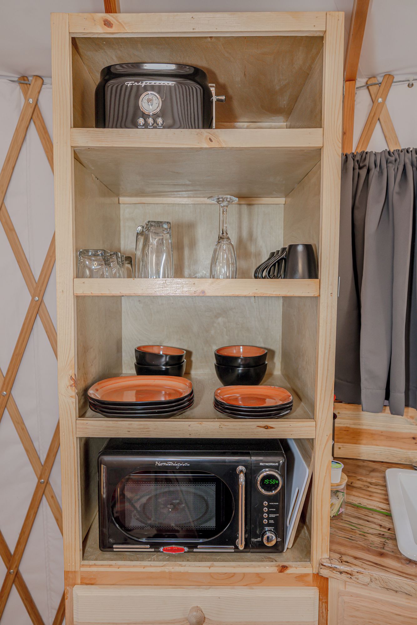 A wooden shelf unit with a retrofitted microwave, stacked plates, and a glass pitcher.