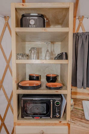 Un estante de madera con un microondas adaptado, platos apilados y una jarra de vidrio.