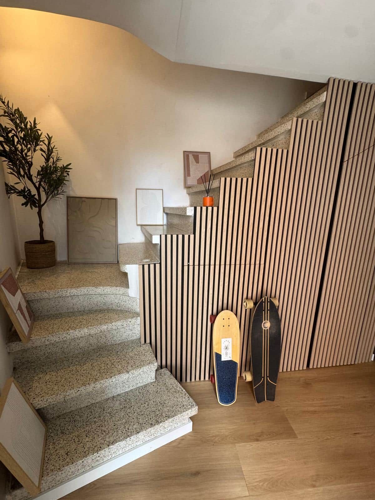 Un escalier avec des finitions en bois, une plante en pot et deux planches de skate appuyées contre un mur composé de lattes de bois verticales.