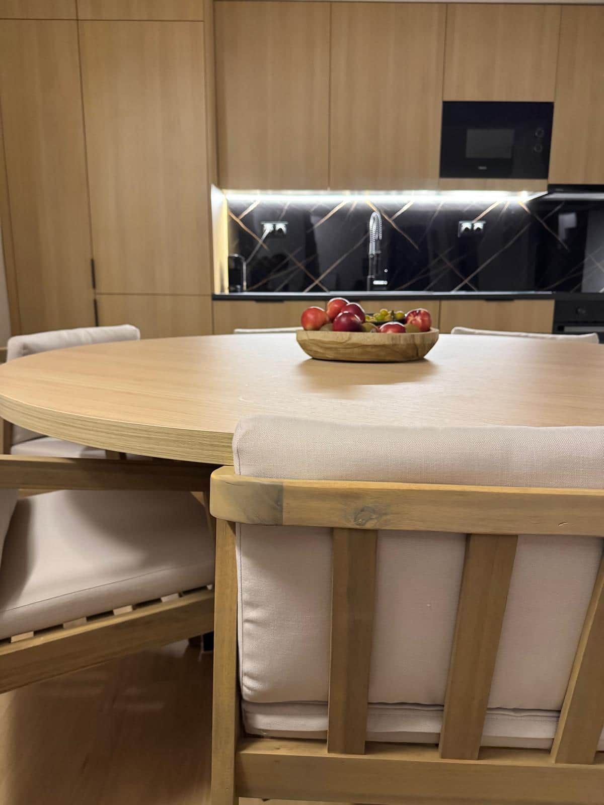 Une cuisine moderne avec une table de salle à manger ronde, des chaises en bois et un saladier de fruits posé sur la table.