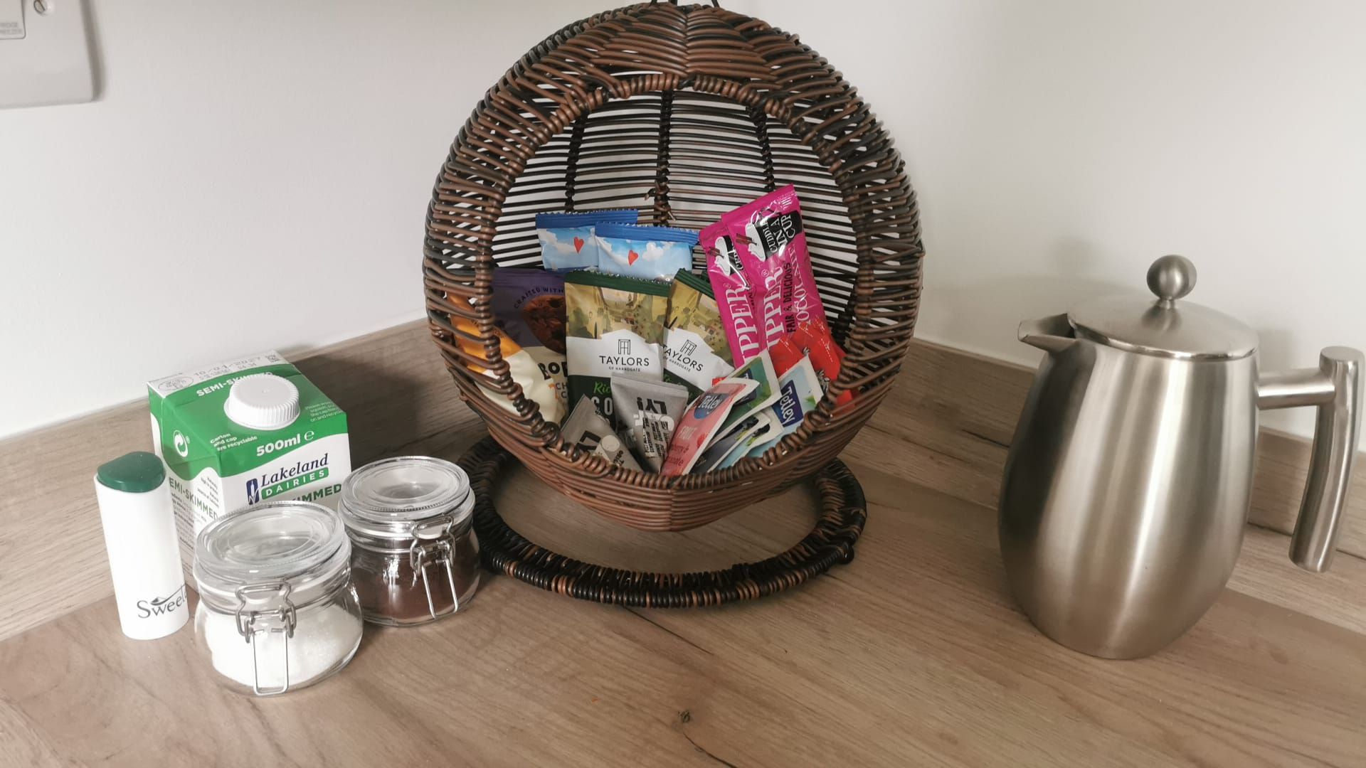 Een rieten mand gevuld met tijdschriften en post, een melkverpakking, twee potten en een roestvrijstalen waterkoker op een houten oppervlak.