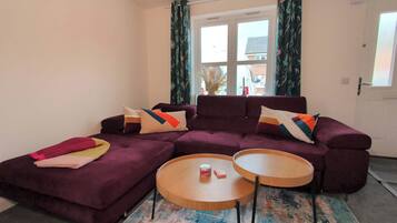 Ein Wohnzimmer mit einem violetten Ecksofa, zwei runden Holztischen und einem farbenfrohen Teppich.