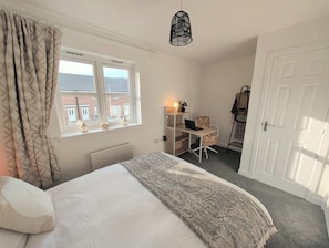A bedroom with a bed, a desk, and a chair. There is a window with curtains, a radiator, and a door. - Stylish 2-Bed Home, Workspaces & Parking (Edinburgh)