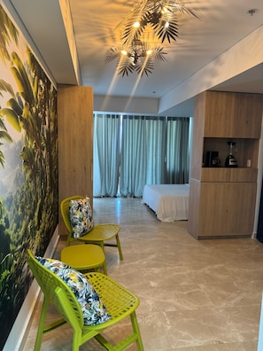 A hotel room with a bed, a seating area with chairs, a painting of a tropical landscape, and a kitchenette. - Enjoyable 1-bedroom hotel , ocean view with AC in charming Cartagena de Indias (Cartagena de Indias)