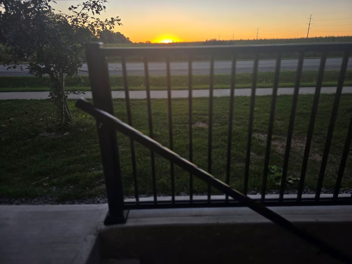 A view of a sunset over a grassy field with a metal fence in the foreground.