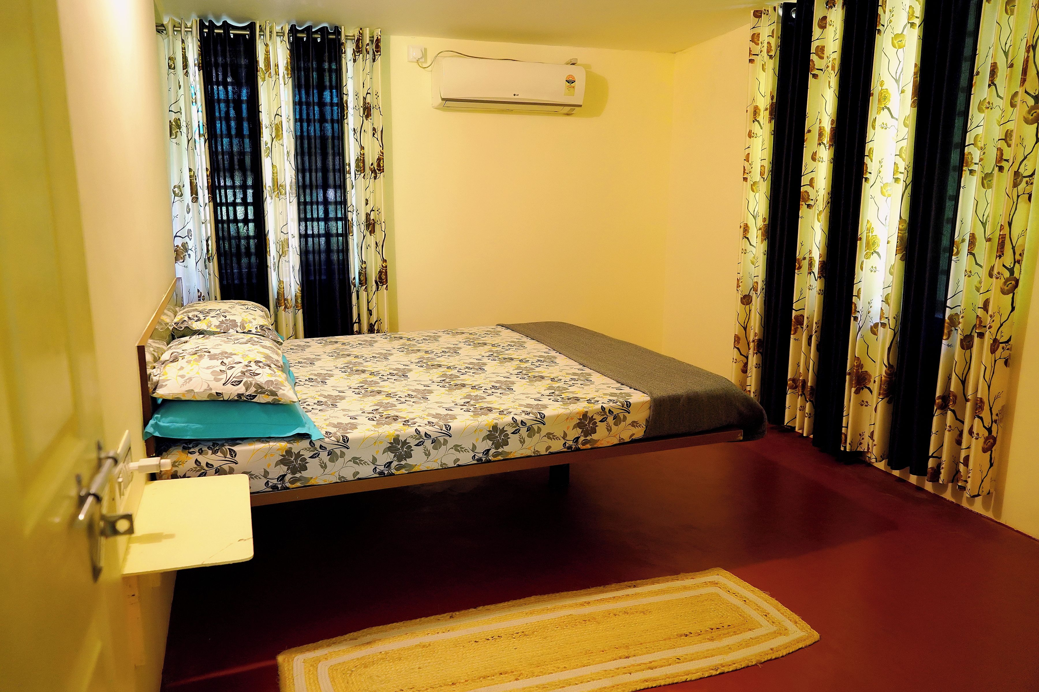 A small, single-bed room with a bed, a bedside table, a wall-mounted air conditioner, and curtains with a floral pattern.