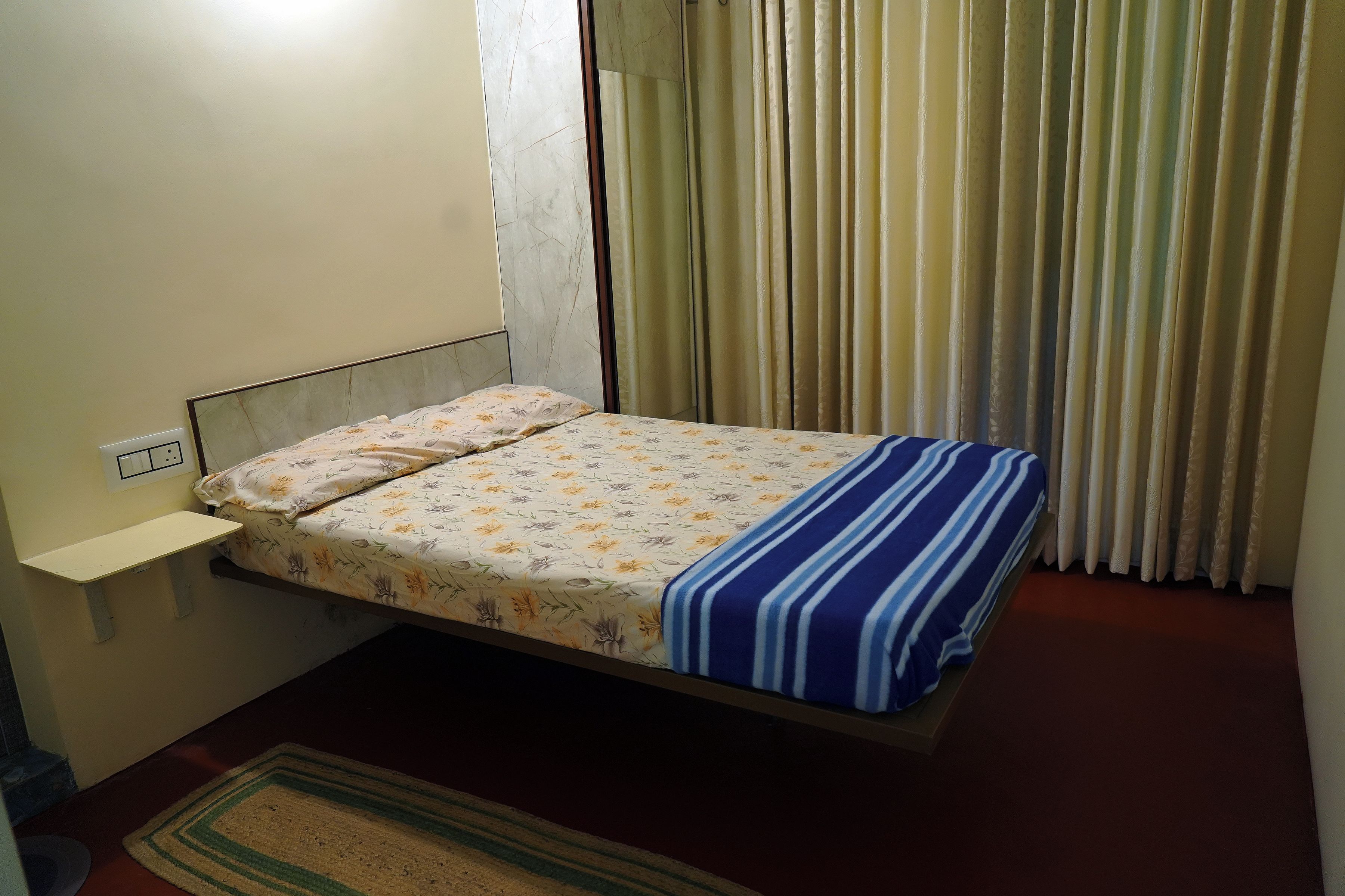 Un lit simple avec une couvre-lit à motifs floraux et une couverture bleue à rayures, une table de chevet et une fenêtre avec des rideaux.