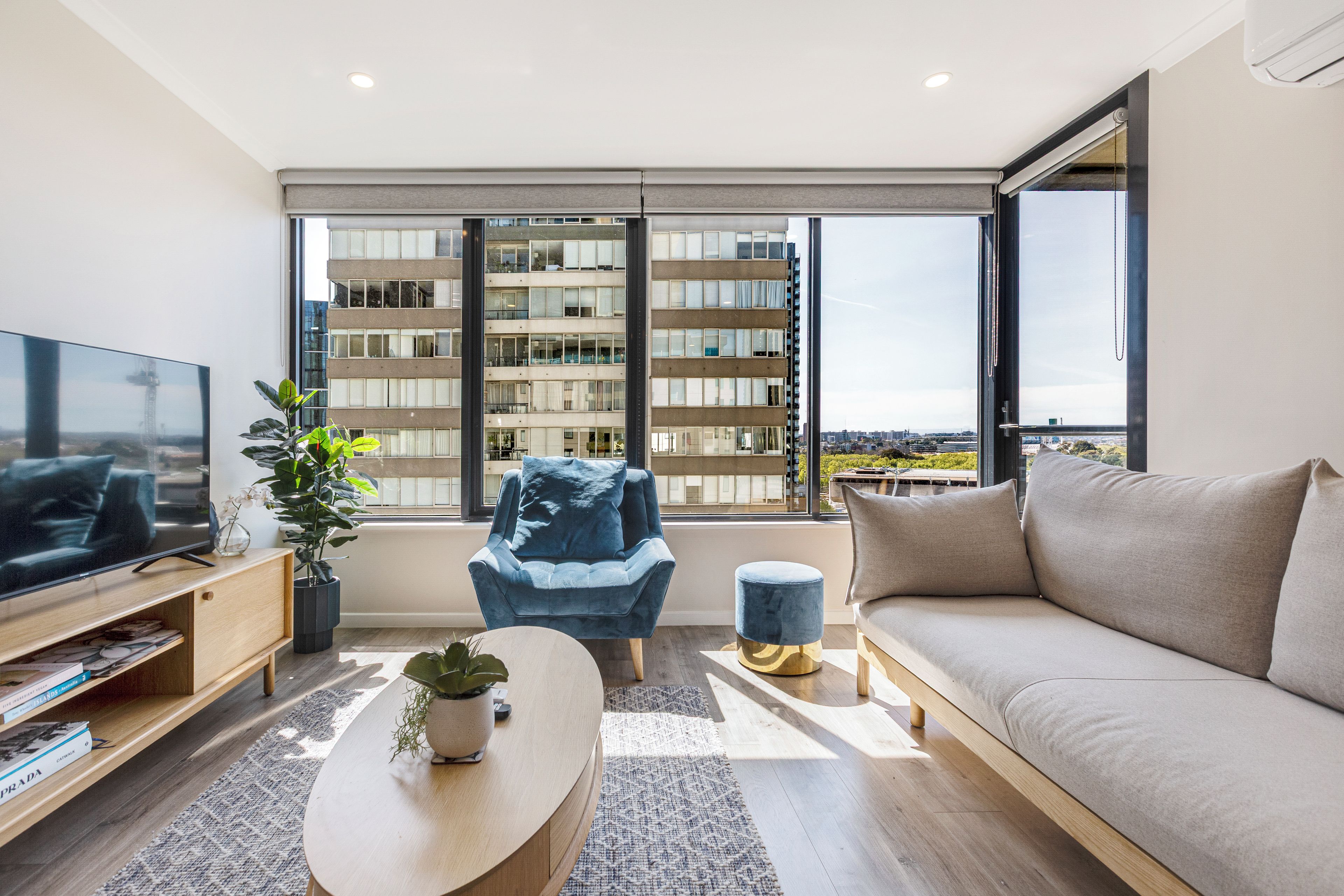 A modern living room with a flat-screen TV, a sofa, a blue armchair, a coffee table, and a large window offering a view of a cityscape.