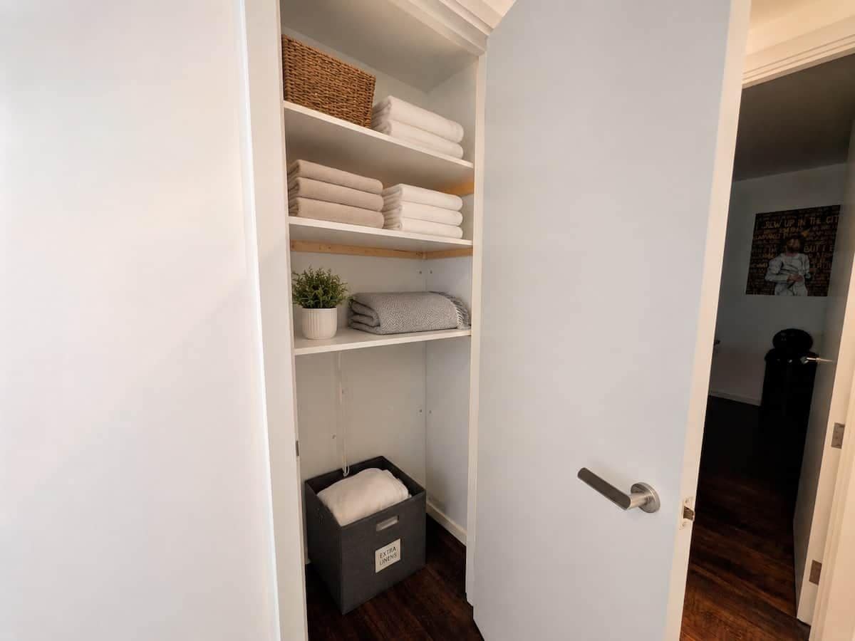 Un dressing avec des étagères, un panier, des serviettes pliées, une plante en pot et une boîte de rangement étiquetée « Linge supplémentaire ».