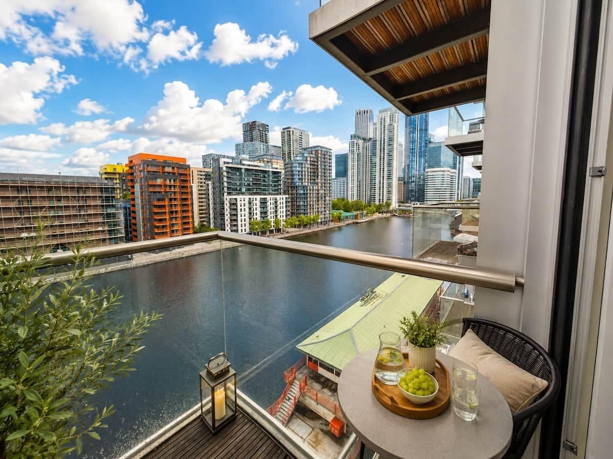 Un balcon avec vue sur une rivière et les bâtiments de la ville, une table avec une boisson et des fruits, et une plante en pot.