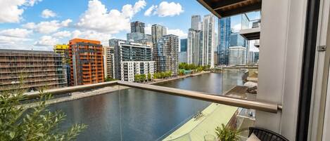 A balcony with a view of a river and city skyline, a table with a drink and fruits, and a potted plant.