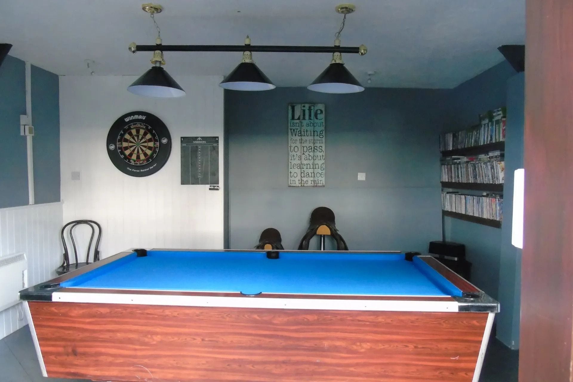 A room with a pool table, dartboards, a poster, and a bookshelf.