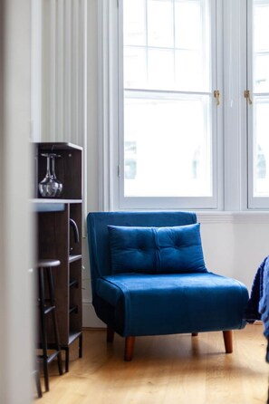 A room with a blue sofa, a wooden floor, a window with blinds, and a dark-colored cabinet.