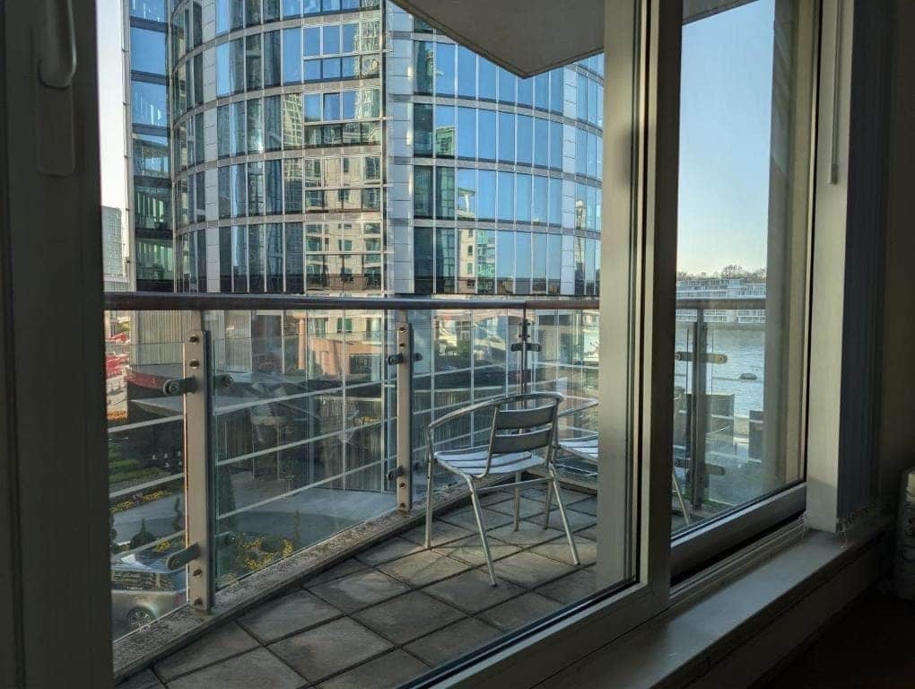 A modern building with a glass facade, a balcony with a chair, and a view of a body of water.