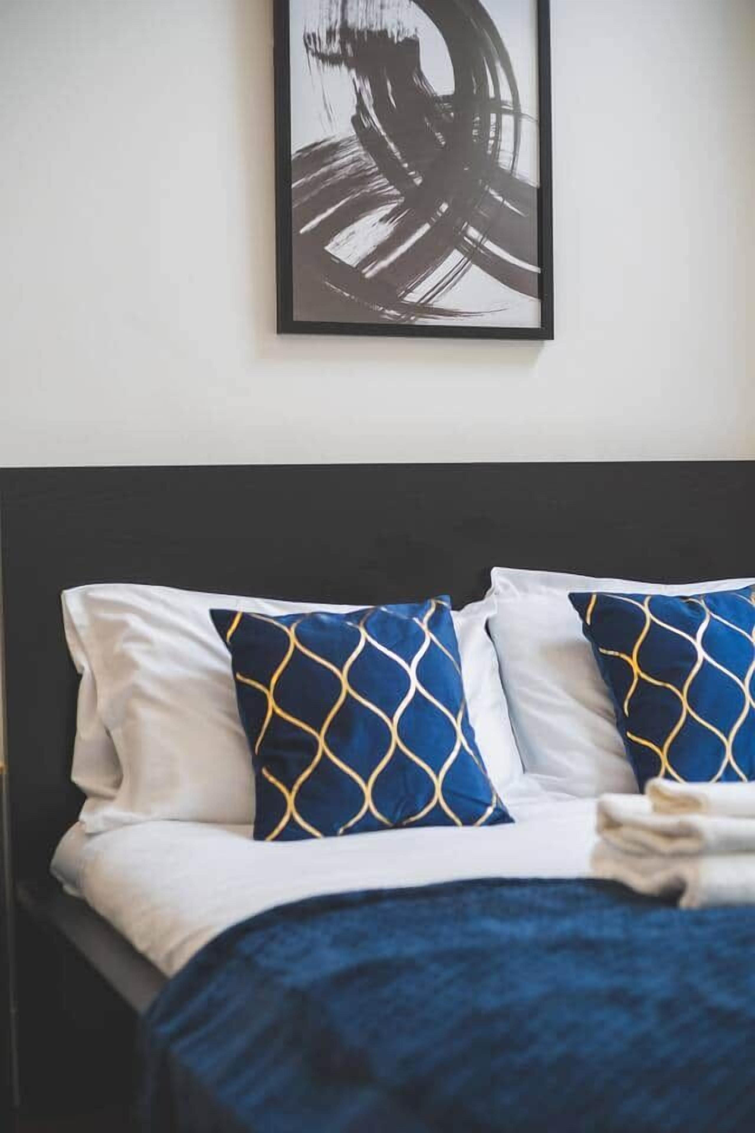 A bed with white and blue pillows, a dark headboard, and a framed abstract painting on the wall.