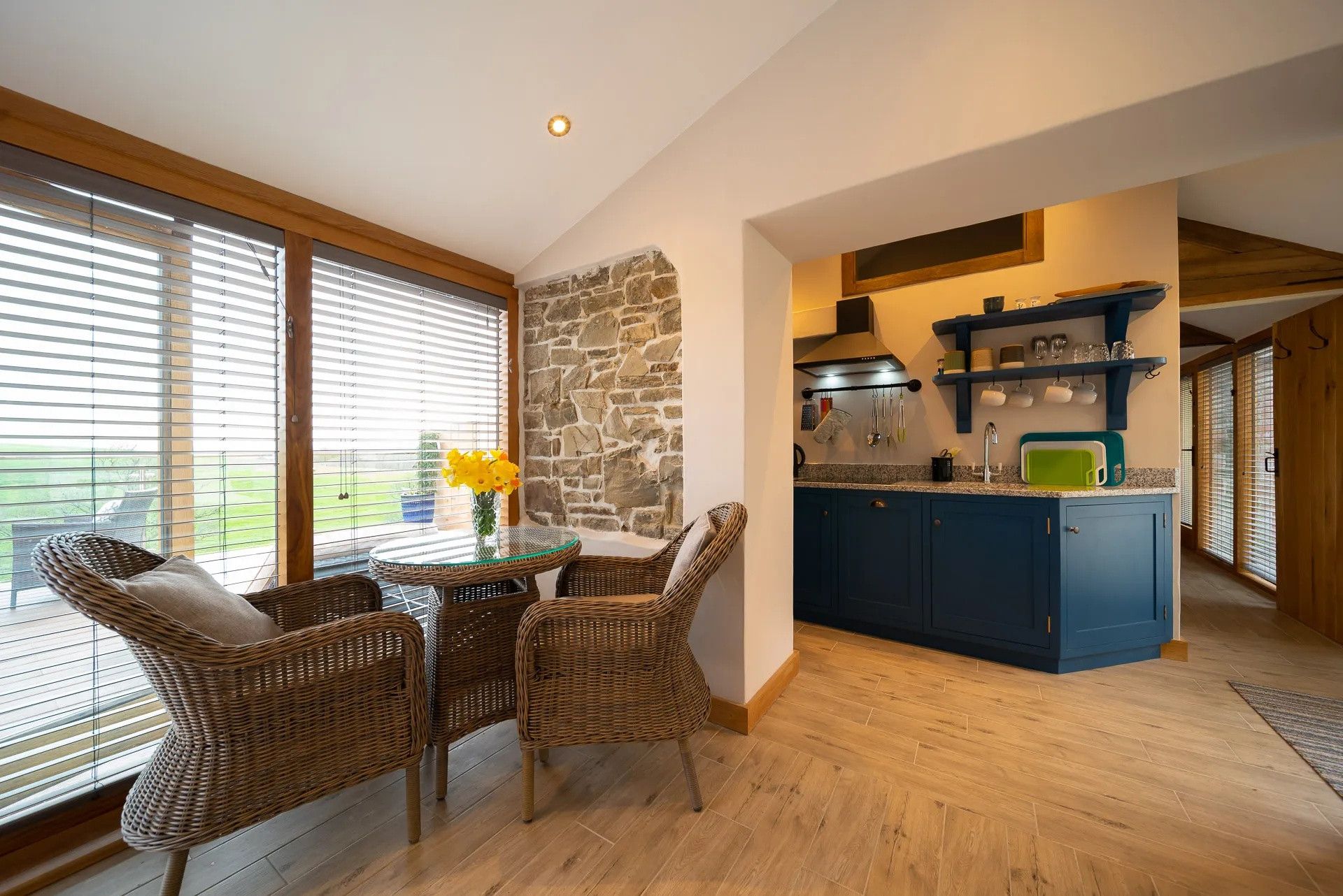 A kitchen with a stone wall, blue cabinets, and a dining area with wicker chairs and a glass table.