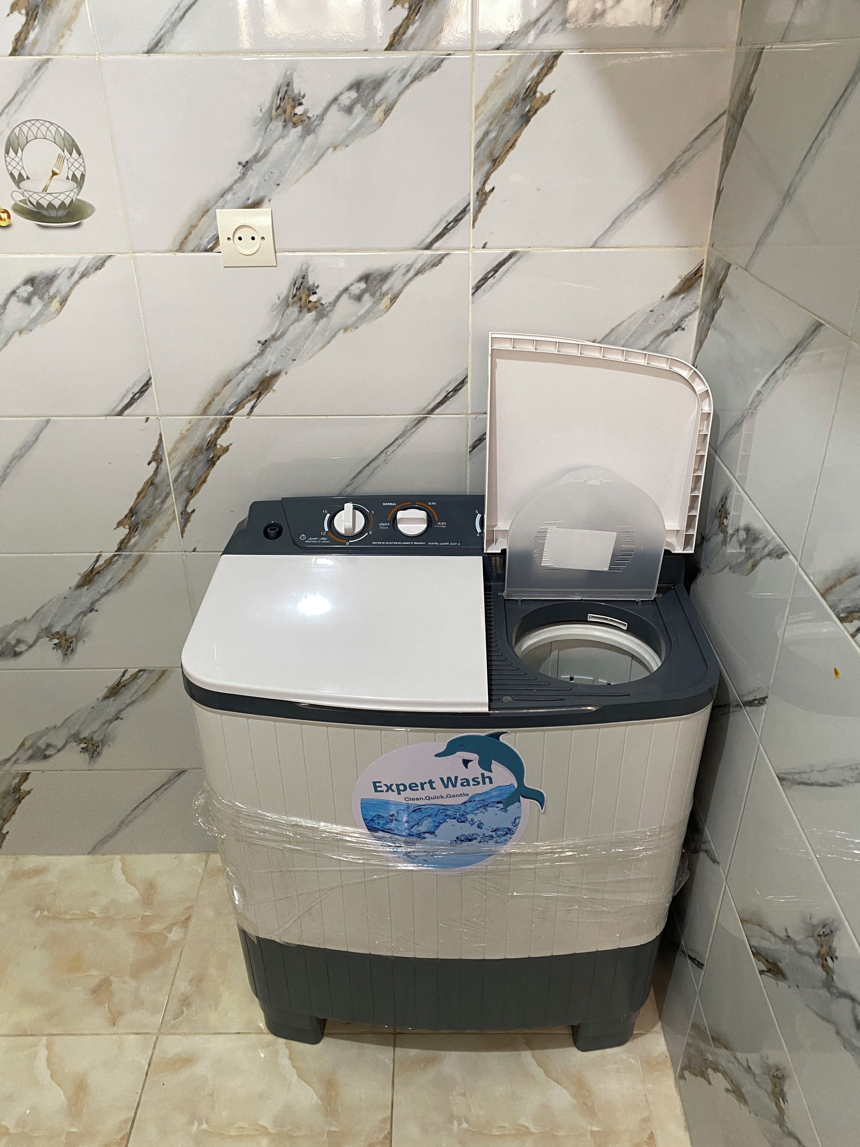 A compact, white washing machine with a front-loading door, placed in a corner against a wall with a marble-like tile pattern.