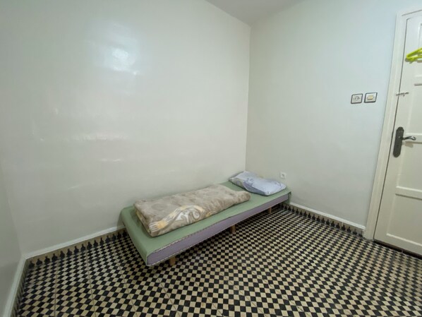 Una stanza piccola con pavimento a scacchi, un letto singolo con cuscini e una porta con maniglia.