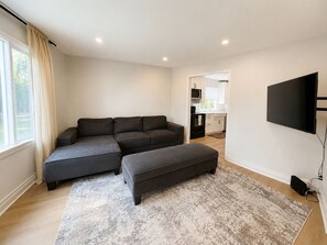 A modern living room with a sectional sofa, a flat-screen TV, and a kitchen area visible through an open doorway. - Modern 4BR Home Near Downtown Winnipeg (Winnipeg)