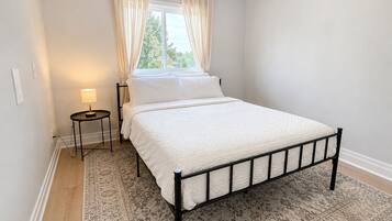 A bedroom with a bed, a bedside table with a lamp, a window with curtains, and a patterned rug.