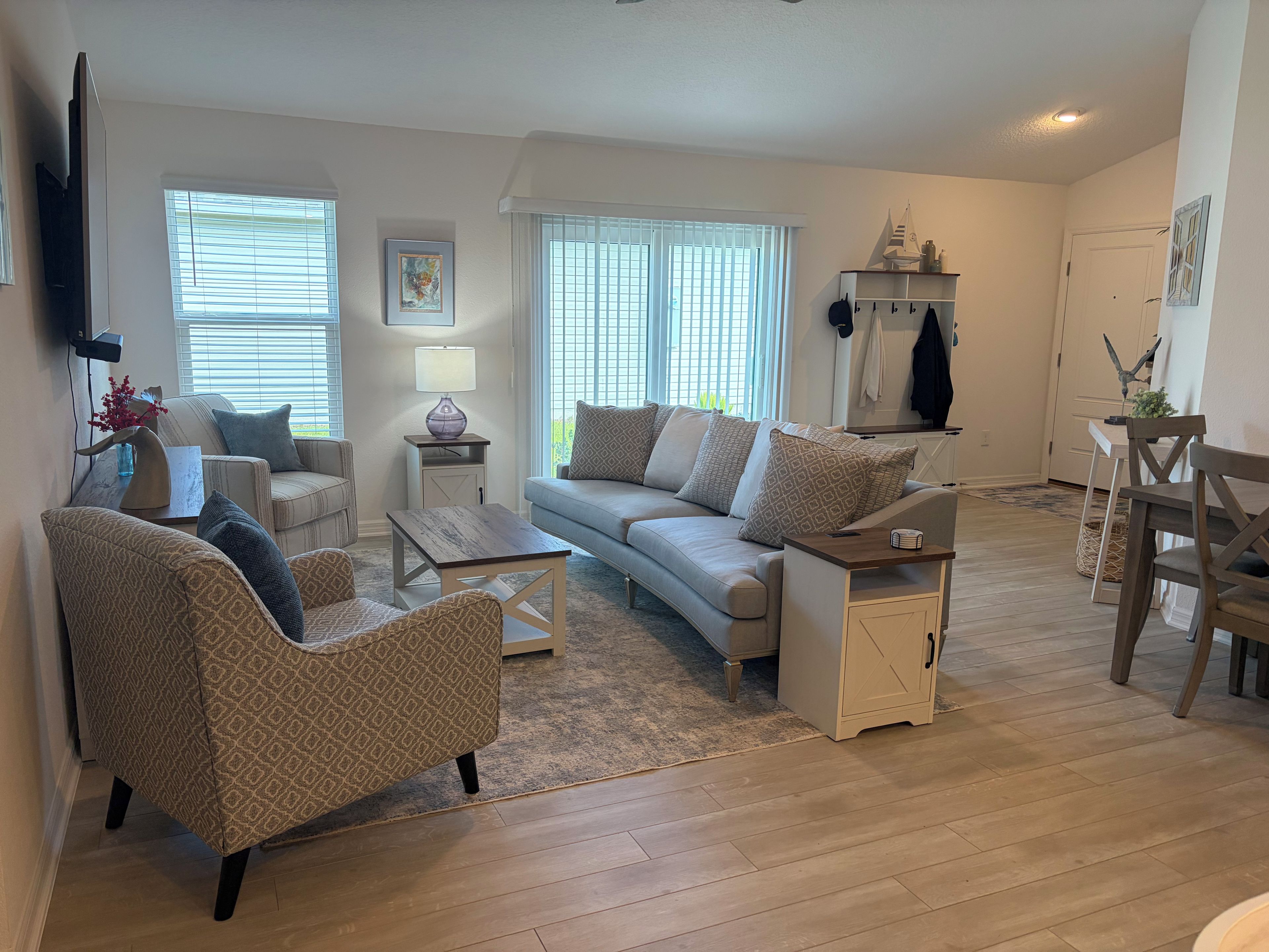 Una sala de estar con un sofá, sillones, una mesa de centro y un televisor. Hay ventanas con persianas, una lámpara y una estantería decorativa.