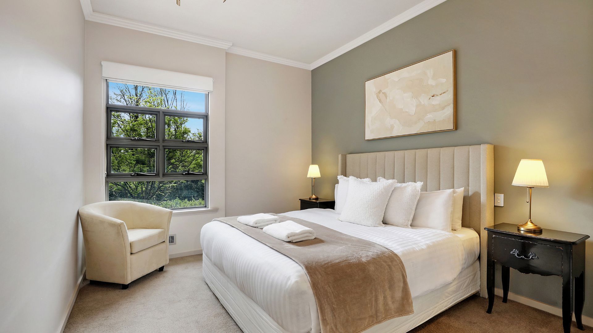 Habitación de hotel con una cama grande, dos lámparas de noche, una silla y una ventana con vista a árboles.