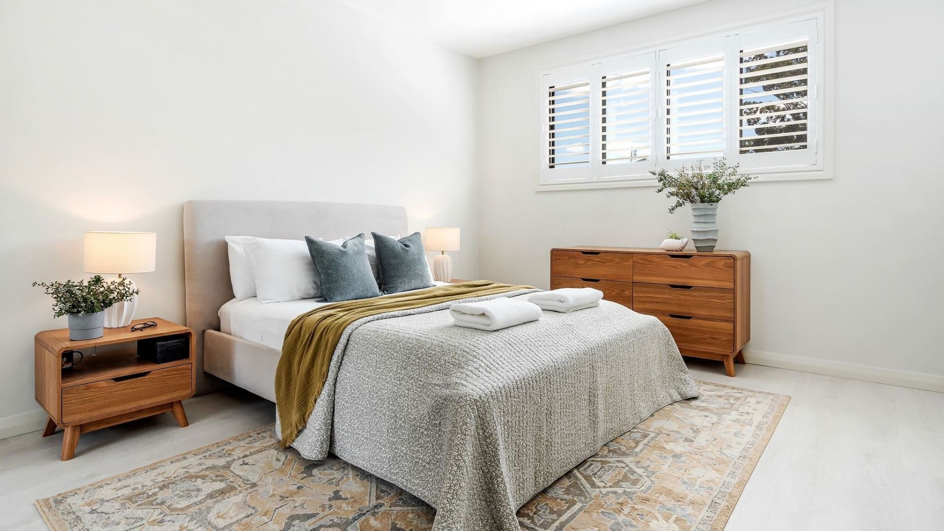 Een slaapkamer met een bed, nachtkastjes, een dressoir, een raam met zonwering en een kamerbreed tapijt met dessin.