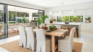 A modern kitchen with white cabinets, a dining area with a large table and chairs, and a view of the outdoors. - Palma – amazing coastal oasis at Callala Beach in beautiful Jervis Bay (Callala Beach)