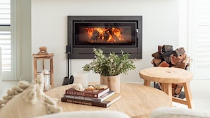 A cozy living room with a fireplace, a round wooden table, books, a potted plant, and a small stool. - Palma – amazing coastal oasis at Callala Beach in beautiful Jervis Bay (Callala Beach)