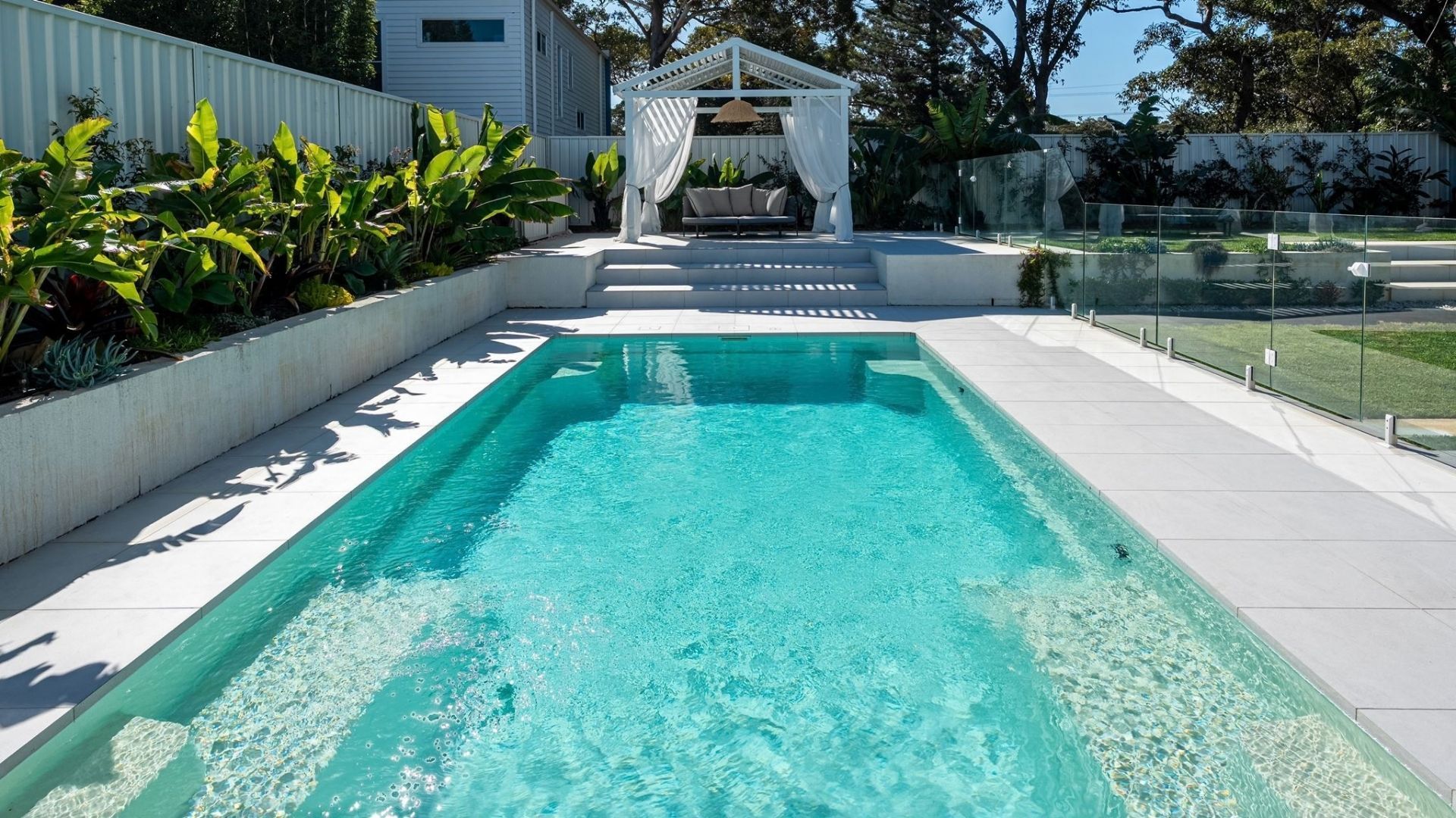 A rectangular swimming pool with clear blue water, surrounded by a white deck and a covered seating area with a canopy.