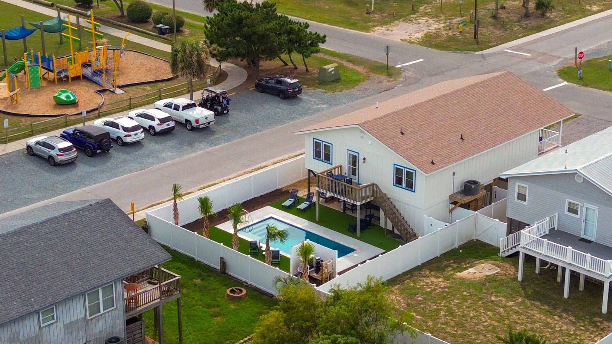 A residential area with a house featuring a swimming pool, a fenced yard, and a playground in the background.