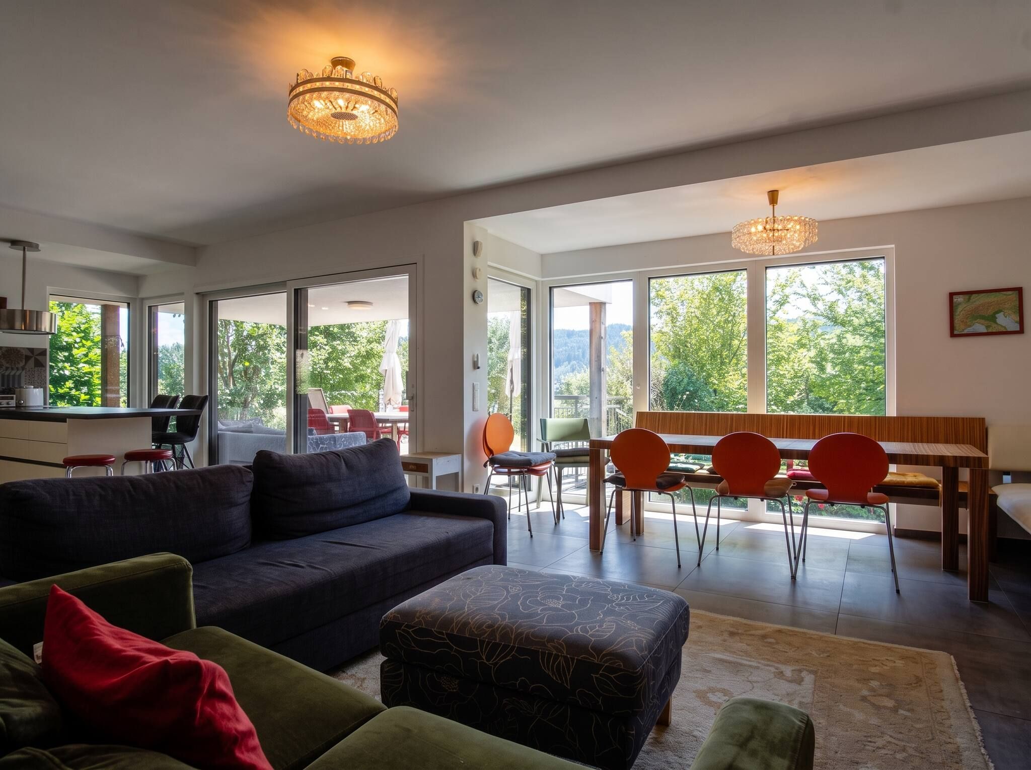 Ein großzügiger Wohnbereich mit einer Couch, Hocker und einem Esstisch mit Stühlen. Große Fenster bieten einen Ausblick ins Freie.