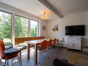 A modern interior with a dining area, seating, a television, and a view of greenery outside. - Lichtblick Garden - Near the Lake by WÖRTHWHILE (Seeboden)