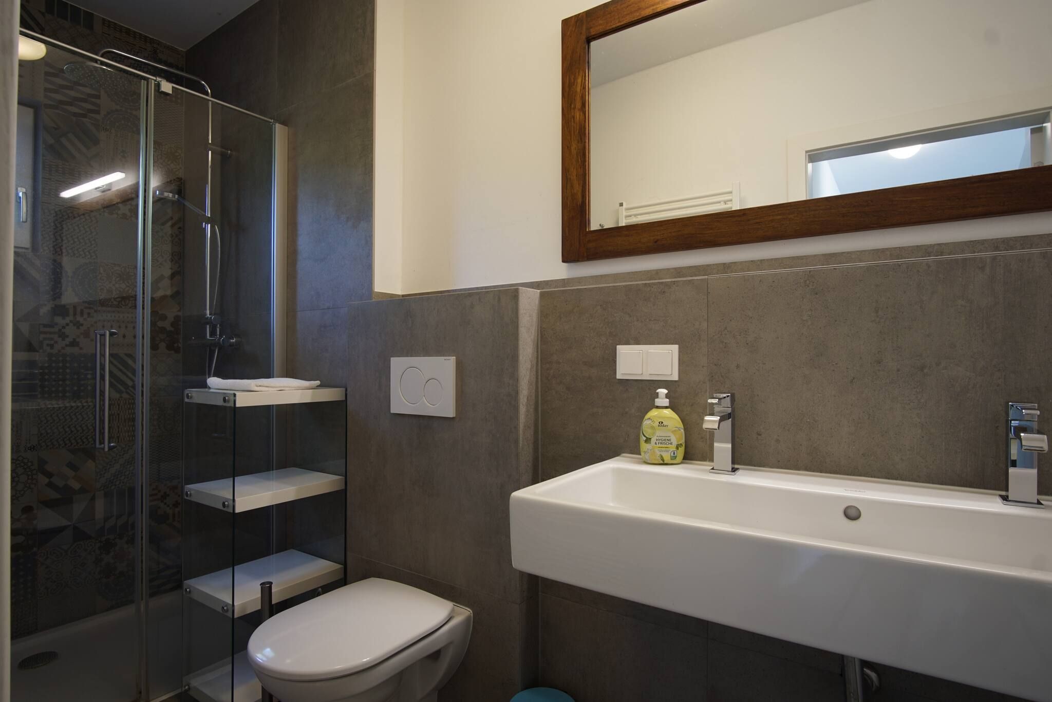 Ein modernes Badezimmer mit Duschkabine aus Glas, einer weißen Toilette, einem großen Waschbecken mit Spiegel darüber und einer Handseifenflasche am Waschbecken.