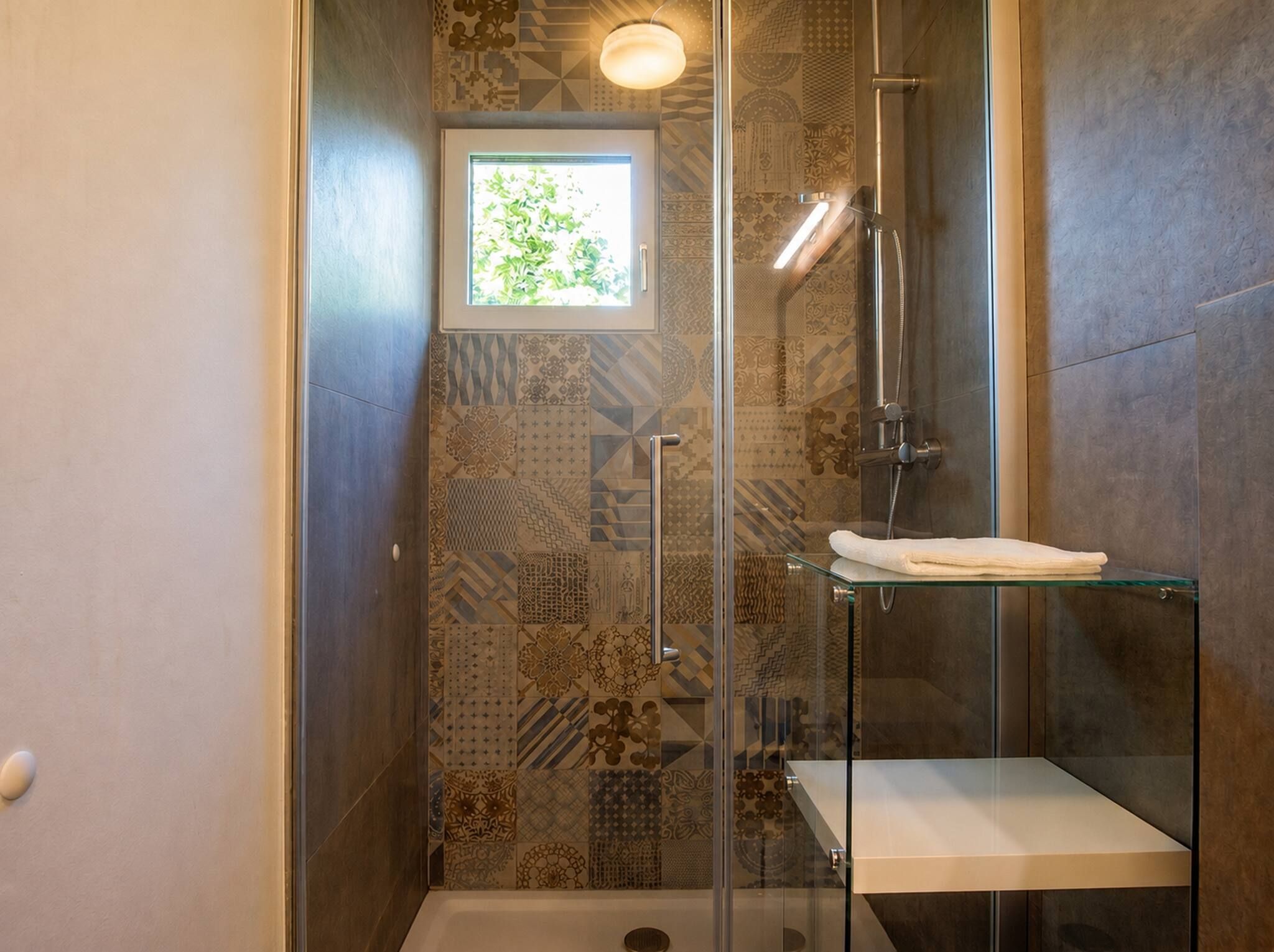 A shower with a glass door, a small window, and a towel on a glass shelf.