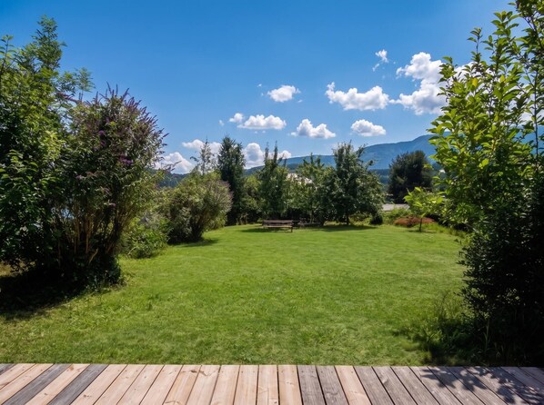 A well-maintained grassy lawn with a wooden deck in the foreground, surrounded by dense shrubbery and trees. - Lichtblick Garden - Near the Lake by WÖRTHWHILE (Seeboden)