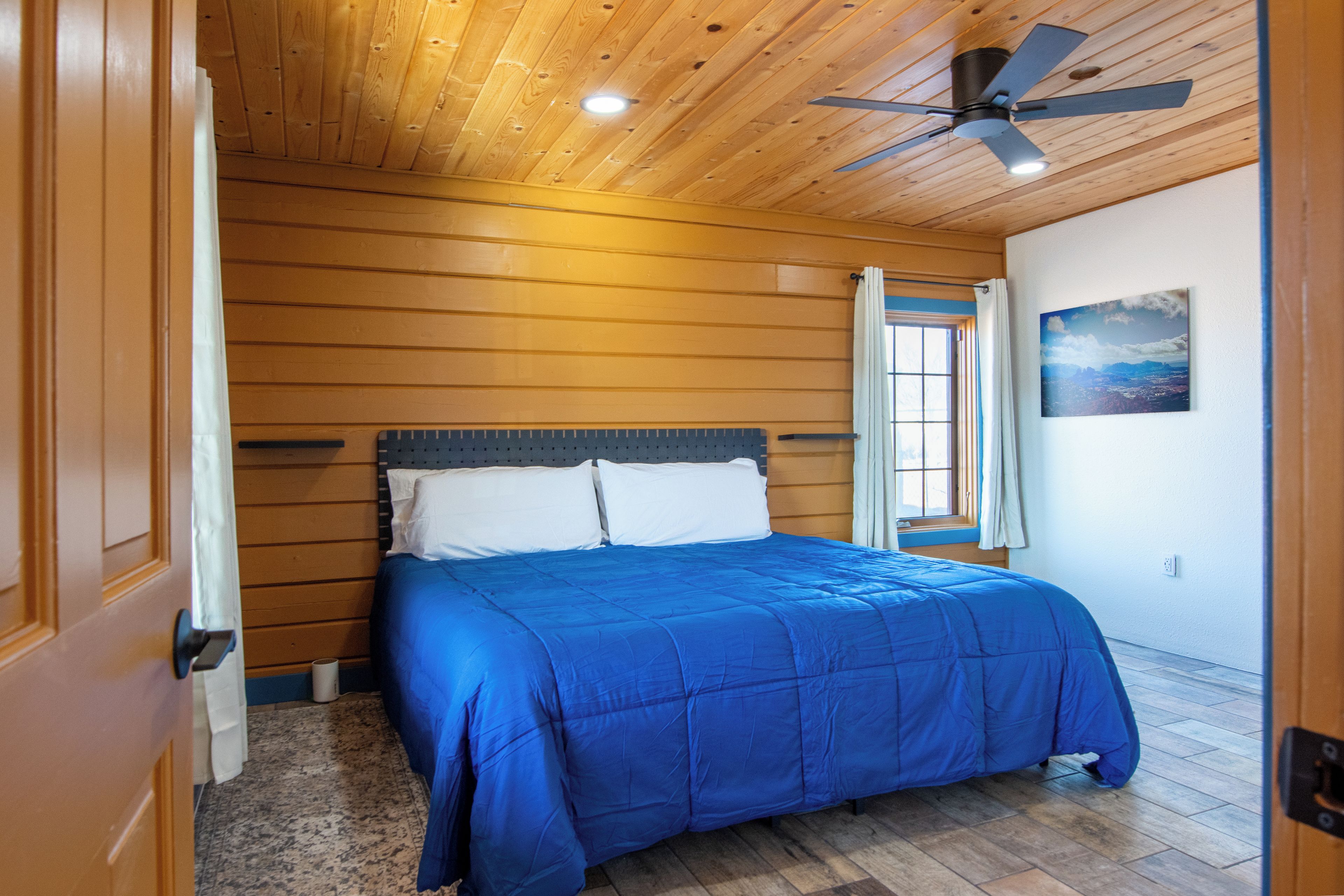 A bedroom with a wooden ceiling, a bed with a blue bedspread, a window with curtains, and a painting on the wall.