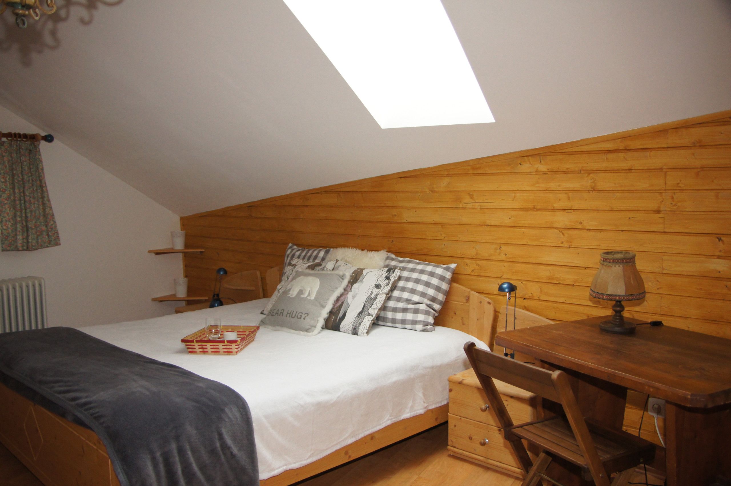 A cozy bedroom with a sloped ceiling, a wooden headboard, a bed with multiple pillows, a bedside table with a lamp, and a small wooden dresser.