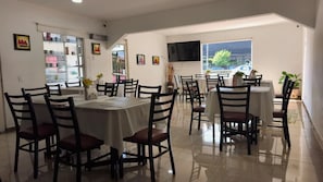 Un comedor con varias mesas y sillas, un televisor montado en la pared y vistas al exterior a través de una ventana.