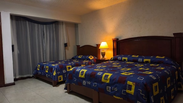 Habitación de hotel con dos camas, una mesita de noche con lámpara, una ventana con cortinas y suelo de baldosas.
