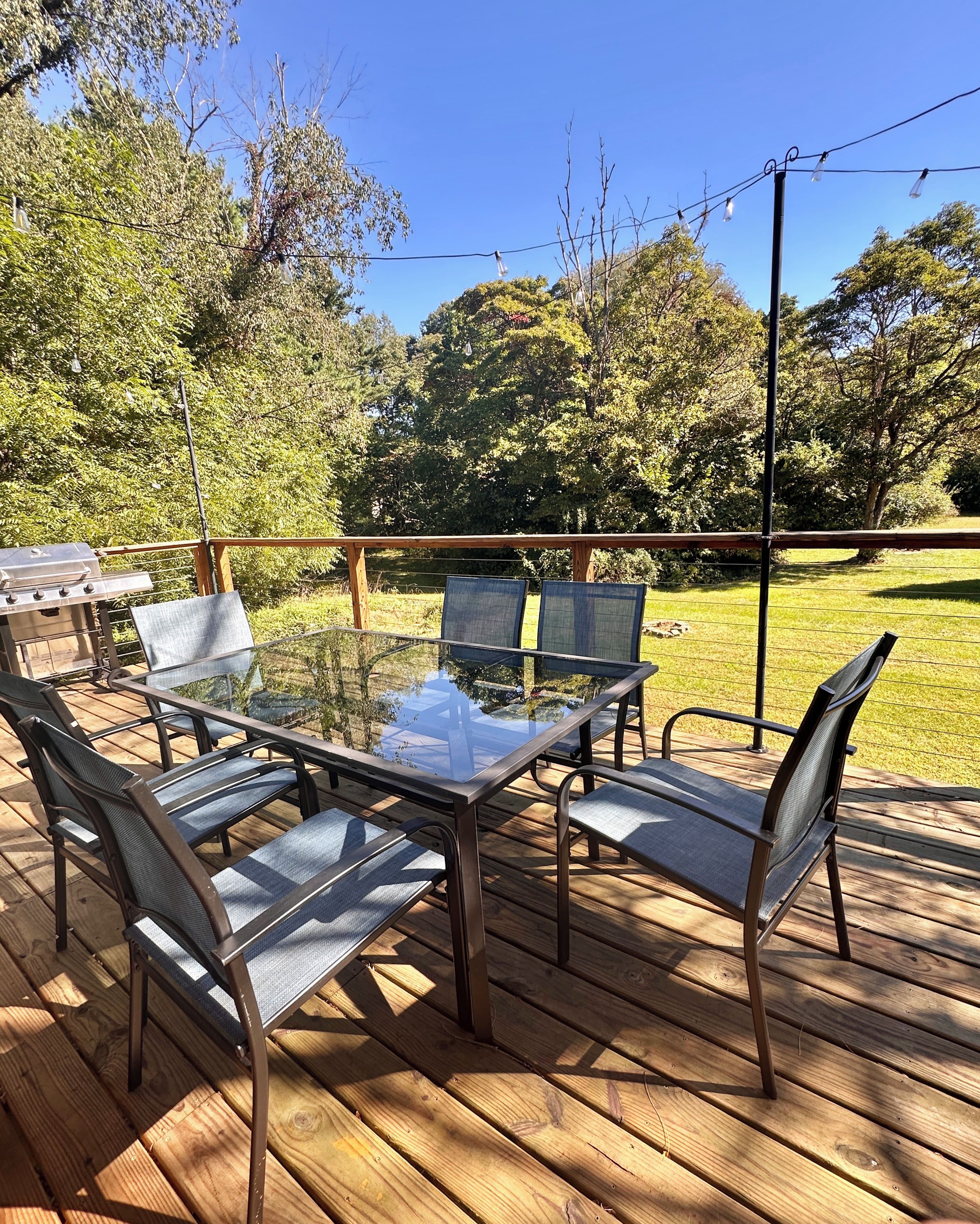 A wooden deck with a glass-top table and metal chairs, surrounded by a wooden fence and trees.