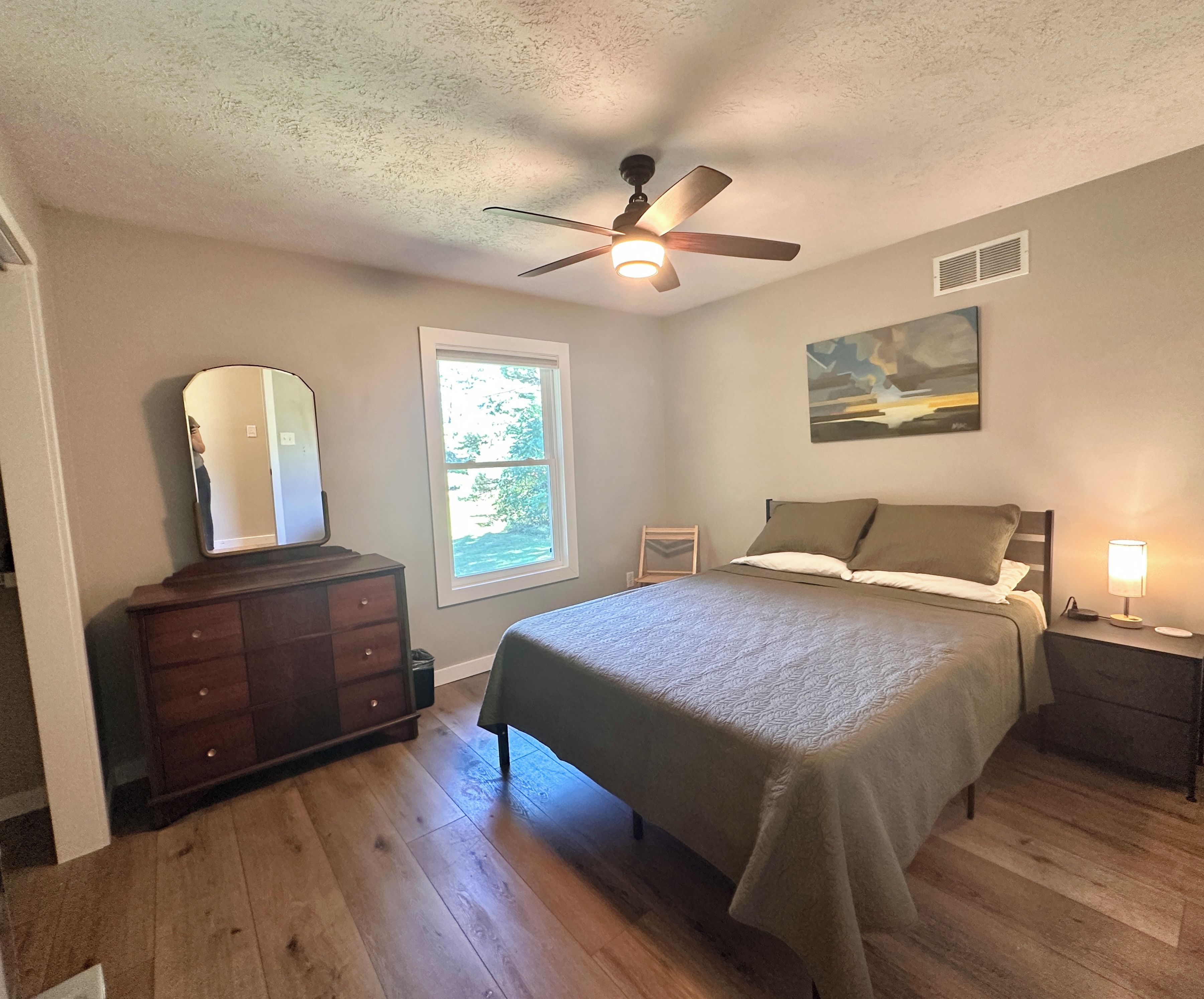 A bedroom with a bed, dresser, ceiling fan, window, and artwork on the wall.