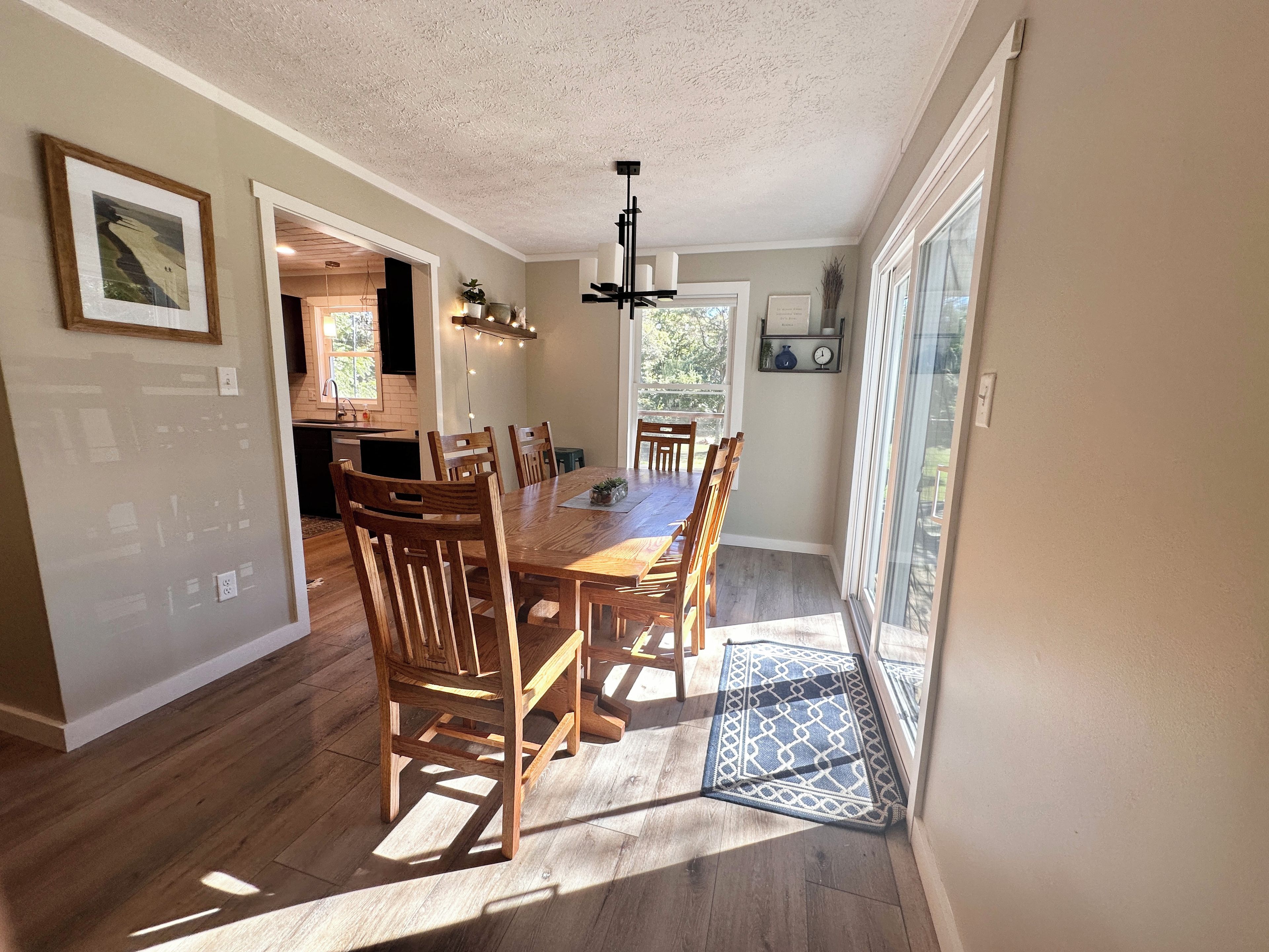 A dining area with a wooden table and chairs, a kitchen in the background, and a view through a glass door.
