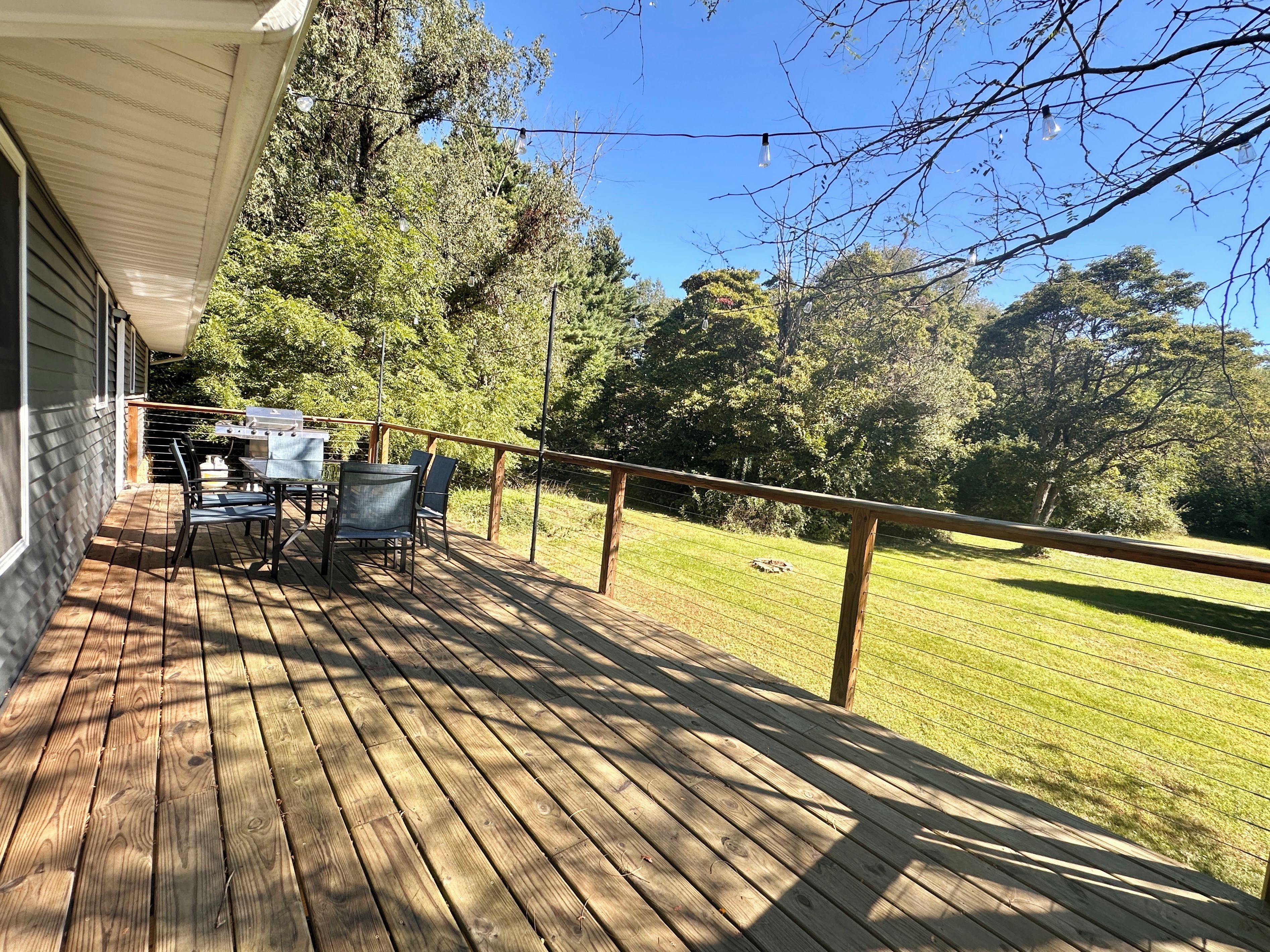 Une terrasse en bois avec des meubles d’extérieur, un barbecue et une vue sur une pelouse verdoyante bordée d’arbres.