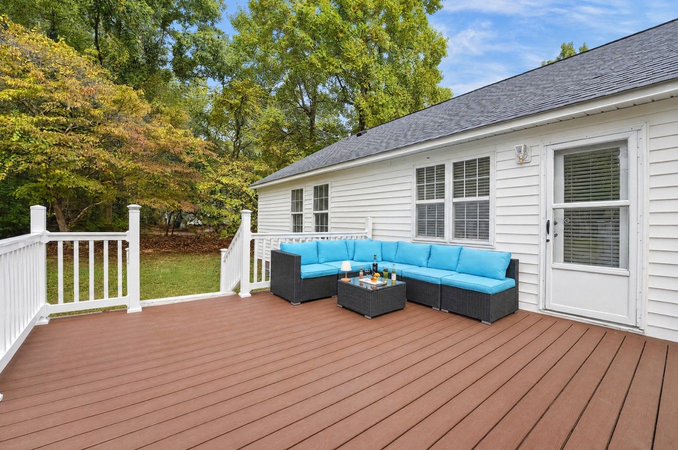 Une terrasse couverte avec un canapé bleu et une table basse, des rampes blanches et une maison aux murs recouverts de bardage blanc et dotée d’un toit gris.