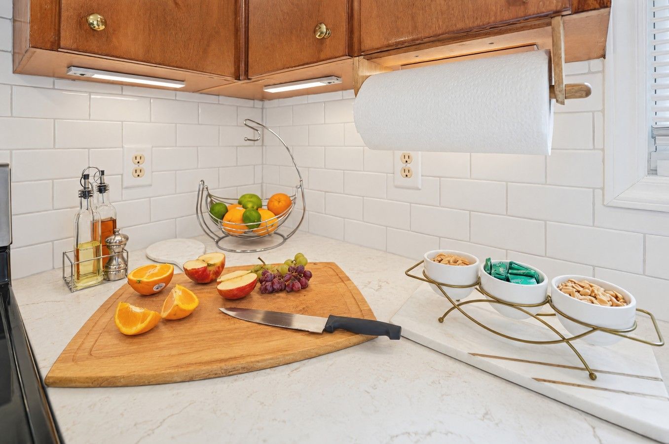 Une cuisine avec des carreaux de céramique blancs disposés en motif métro, des armoires en bois, une planche à découper en bois avec des fruits et un couteau, et un distributeur d’essuie-tout.