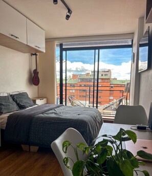 Ein modernes Schlafzimmer mit Bett, einer Gitarre, einem Schreibtisch mit Laptop und einem Balkon mit Blick auf Gebäude.