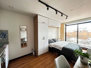 A bedroom with a bed, a wardrobe, a desk, a chair, a potted plant, and a window with a view of buildings. - New Listing (Bogotá)